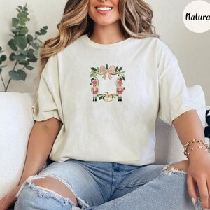 Puede incluir: Camiseta color crema con un dise&ntilde;o de corona festiva con lazo rosa, hojas verdes y dos figuras de soldados de juguete. La palabra "Natural" est&aacute; impresa en la parte superior derecha. La camiseta es de material suave.