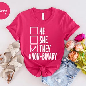 May include: A magenta t-shirt with the text "HE SHE THEY #NON-BINARY" in white. The shirt has a crew neck and rolled-up sleeves. The image also includes a pair of beige sandals, denim shorts, and a bouquet of flowers.