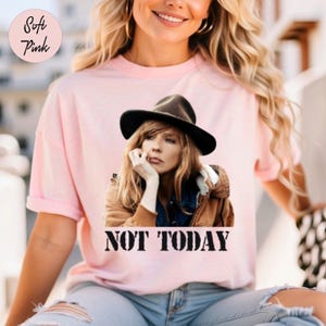 Peut inclure: T-shirt rose clair avec un imprimé d'une femme portant un chapeau et le texte "NOT TODAY" en noir. Le t-shirt a des manches courtes et une coupe ample. Les mots "Soft Pink" sont dans un cercle en haut à gauche.