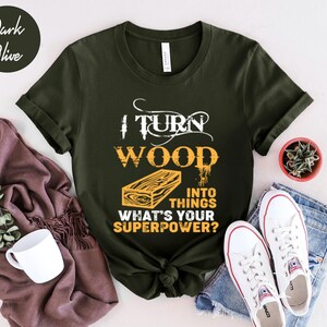 May include: Dark olive green t-shirt with a wood grain design and the text "I turn wood into things. What's your superpower?"