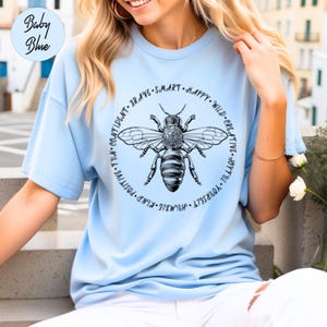 Peut inclure: Un t-shirt bleu clair avec une illustration d'abeille noire et blanche au centre. L'abeille est entourée d'un cercle de texte qui dit : "HUMBLE. KIND. POSITIVE. FUN. CONFIDENT. BRAVE. SMART. HAPPY. WILD. CREATIVE. JOYFUL. YOURSELF."