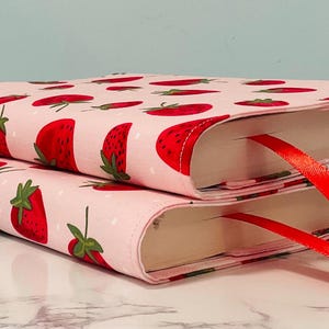 Puede incluir: Dos cubiertas de libros rosas con un patrón de fresas y marcadores de cinta roja. Las cubiertas están apiladas una encima de la otra.