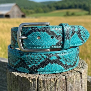 May include: A turquoise and black snakeskin-patterned belt with a silver buckle. The belt is rolled up and resting on a wooden post. The background features a field, a barn, and a blue sky.