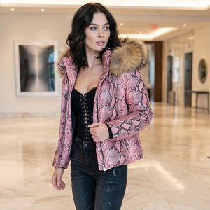 May include: A pink snakeskin-effect jacket with a fur-trimmed hood. The jacket is zipped and worn over a black top and dark jeans. The model is wearing black boots with a matching snakeskin pattern.