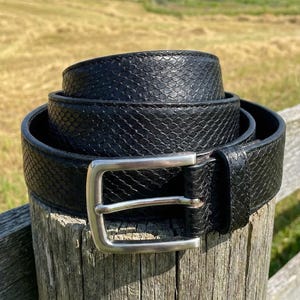 May include: A black belt with a snakeskin texture and a silver buckle is coiled on a wooden post. The belt is the focal point, with a rural landscape in the background, including a red barn and rolling hills under a blue sky.
