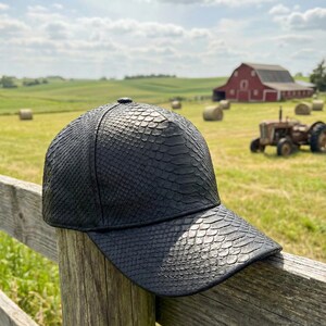 Può includere: Un cappellino da baseball nero con un motivo strutturato simile alla pelle di serpente. Il cappellino è appoggiato a un paletto di legno. Lo sfondo mostra un paesaggio rurale con un fienile rosso, balle di fieno e un trattore.