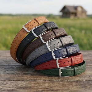 May include: A selection of leather belts in shades of tan, black, brown, grey, navy, red, and green. Each belt has a silver buckle and a textured finish, possibly ostrich leather. The belts are displayed in a fanned arrangement on a wooden surface.