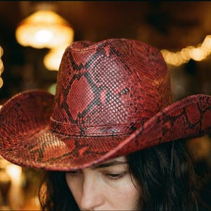 Könnte beinhalten: Ein roter Cowboyhut mit Schlangenmuster. Der Hut hat eine breite Krempe und eine klassische Cowboyhutform. Der Hut besteht aus einem strukturierten Material und hat eine dunkelbraune Einfassung. Ein auffälliges Modeaccessoire.