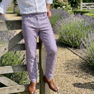 Può includere: Pantaloni color lavanda con motivo a pelle di serpente. I pantaloni sono abbinati a mocassini in pelle marrone. La persona è appoggiata a una staccionata di legno, con un giardino sullo sfondo.