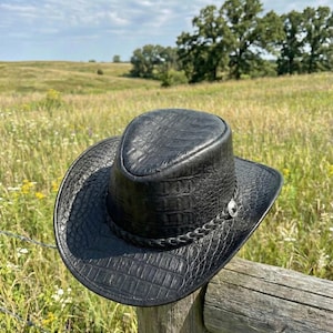 May include: A black cowboy hat with a textured, crocodile-like pattern. The hat has a braided band and a silver-coloured button. It rests on a wooden fence post in a field with a blue sky and trees in the background.