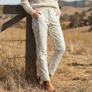 Puede incluir: Pantalones con estampado de serpiente color crema con pernera recta. Los pantalones tienen una paleta de colores neutros de blanco, gris y beige. Los pantalones se combinan con botas de cuero marrones.