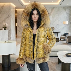 Peut inclure: Une veste matelassée jaune à motif peau de serpent avec une capuche et des poignets bordés de fourrure. La veste est zippée et portée par une personne. L'arrière-plan est un intérieur moderne avec des murs en marbre et un canapé blanc.