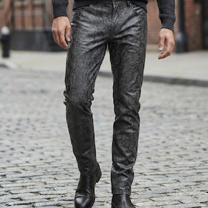 Op de afbeelding: Een persoon draagt een zwarte broek met slangenpatroon en zwarte leren laarzen. De broek heeft een textuur, schubachtig ontwerp. De persoon staat op een geplaveide straat en draagt een donkergrijze trui.