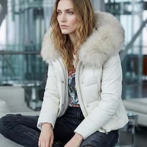 Puede incluir: Chaqueta acolchada blanca con capucha ribeteada de piel. La persona lleva una camiseta gráfica y vaqueros oscuros. La chaqueta tiene cierre de cremallera. La persona lleva botas negras. Abrigo de invierno.