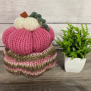 May include: A stack of three knitted pumpkins, the top one is white with a green stem, the middle one is pink, and the bottom one is brown and white.