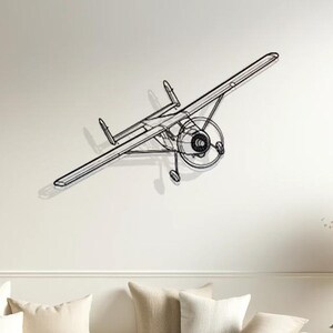 Puede incluir: Una escultura de alambre negro de un avión pequeño colgando de una pared blanca sobre un sofá blanco con cojines beige. El sofá está en una sala de estar con suelo de madera y una mesa auxiliar con un jarrón de flores secas.