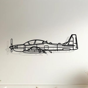 Può includere: Un contorno di aereo in metallo nero montato su una parete bianca sopra un divano bianco con cuscini beige. Il divano si trova in un soggiorno con pavimento in legno e un tavolino con un vaso di fiori secchi.