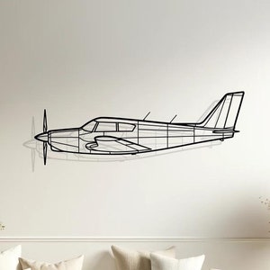Könnte beinhalten: Eine schwarze Flugzeug-Wandskulptur aus Metall vor weißem Hintergrund. Die Skulptur hängt über einem weißen Sofa mit beigen Kissen. Das Sofa steht in einem Wohnzimmer mit Holzboden.