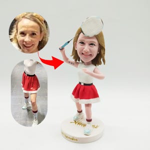 Könnte beinhalten: Eine maßgefertigte Figur einer Person, die Badminton spielt. Die Figur trägt ein weißes Hemd, einen roten Rock mit weißem Besatz und weiße Schuhe. Die Figur hält einen Badminton-Schläger über dem Kopf. Die Figur steht auf einem weißen Sockel mit dem Namen "Alexandra" darauf.