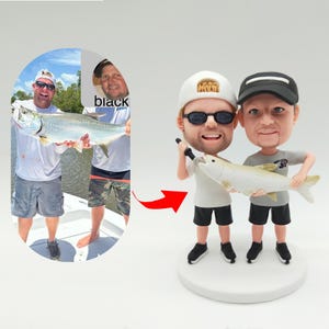 May include: Two custom bobbleheads of men holding fishing rods and fish. The bobbleheads are wearing white shirts, black shorts, and hats. The bobbleheads are standing on a white base.