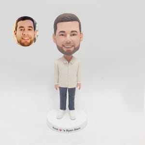 以下が含まれることがあります： ひげと短い茶色の髪をした人のカスタムボブルヘッド人形。人形は白いボタンダウンシャツ、青いジーンズ、白いスニーカーを着用しています。人形の台座には「Knot's Ryan Stern」という文字と赤いハートのシンボルがあります。