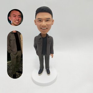 以下が含まれることがあります： グレーのジャケット、黒いシャツ、グレーのパンツを着た人のカスタムボブルヘッドフィギュア。フィギュアは白い台座の上に立っています。