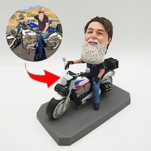 May include: A custom figurine of a person riding a white motorcycle with blue and red accents. The figurine is made of polymer clay and is hand-painted. The person is wearing blue jeans and a dark blue shirt. The motorcycle has a saddlebag on the back.
