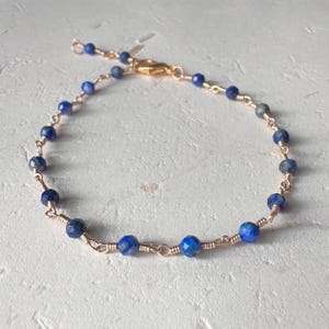 May include: A delicate bracelet featuring small, faceted, deep blue beads strung on a gold-colored wire. The bracelet has a gold-colored clasp and is designed to be adjustable. The beads are evenly spaced, creating a simple yet elegant design.