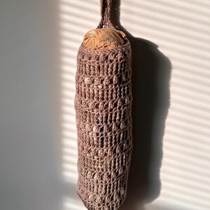 May include: A crocheted plastic bag holder in a muted brown color, hanging from a silver hook. The holder is cylindrical with an open weave design, and a plastic bag is visible inside. The holder is suspended against a white wall with sunlight and shadows.