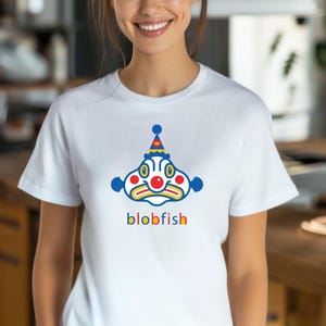 以下が含まれることがあります： 白いTシャツに、青い、黄色い、赤い漫画のピエロの顔とパーティーハット、そして「blob fish」という文字が前面に印刷されています。