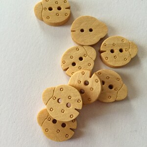 May include: Six wooden buttons shaped like ladybugs with two black dots for eyes and a black dot for a mouth. The buttons are a light brown color.