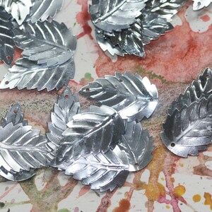 May include: A collection of silver leaf-shaped sequins. The sequins are scattered on a white surface with pink and orange watercolor paint splatters.