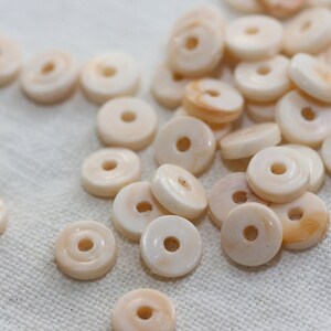 May include: A collection of off-white shell beads with a hole in the center. The beads are round and flat, and they have a slightly iridescent sheen.