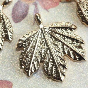 May include: Gold-toned metal leaf charm pendant with a detailed textured surface. The leaf has a pointed tip and a rounded base.