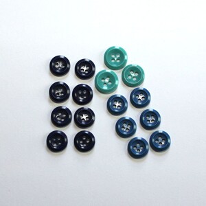 May include: Assortment of blue and teal buttons with four holes in the center. The buttons are arranged in a pattern on a white background.