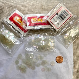 May include: Three packages of iridescent white sequins, each containing 150 pieces. The sequins are 8mm in diameter and are packaged in clear plastic bags with red and white labels that read "SEQUINS" and "No. 1408 8MM 150 PCS."