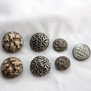 May include: A collection of six antique buttons in various designs. The buttons feature textures like organic patterns, woven designs, and a checkered pattern. Colors include silver, bronze, and a mother-of-pearl effect. The buttons are round and of varying sizes.