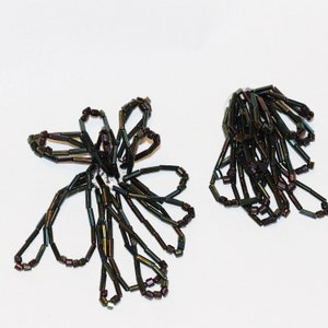 May include: Two black and iridescent beaded flower shapes. The larger flower has a more open shape with petals that curve outward. The smaller flower has a more compact shape with petals that are closer together.
