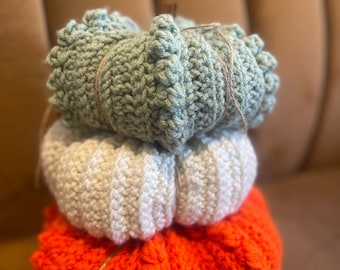 May include: A stack of three crocheted pumpkins in orange, white, and light green. The pumpkins are tied together with twine.