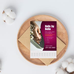 May include: A business card for "Nails by Bella" with a burgundy and white design, featuring a hand with red nail polish reflected in a mirror. The card is on a wooden plate with a brown envelope and cotton accents.