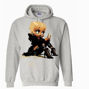 May include: A grey hoodie featuring a cartoon illustration of a woman with blonde hair, wearing a black leather jacket and black trousers. The woman is sitting on the ground and looking at her phone. The text "Kustom & Customs" is printed in gold on the hoodie.