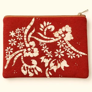 May include: A rectangular zippered pouch in a deep red fabric featuring a cream-colored batik-style floral pattern. The design includes stylized blossoms, leaves, and swirling vines across the front surface, finished with a metallic zipper closure.