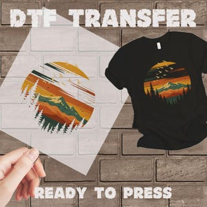 May include: Black t-shirt with a circular graphic of a mountain landscape in shades of orange, yellow, and green. A DTF transfer with the same design is held next to the shirt. The words "DTF TRANSFER" and "READY TO PRESS" are visible.