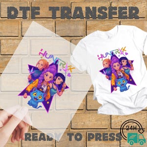 May include: White t-shirt with a colorful graphic featuring three cartoon women and a blue cat, with the word "HUNTRX" in a stylized font. A DTF transfer of the same design is shown next to the shirt, with the words "READY TO PRESS".