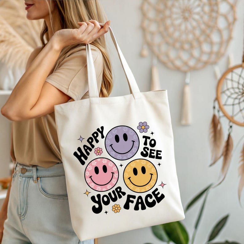 以下が含まれることがあります： 「Happy to See Your Face」の文字と、ピンク、紫、黄色のスマイリーフェイスが3つプリントされた、オフホワイトのキャンバス地のトートバッグ。シンプルなデザインで、2つのハンドルがあり、人が持っています。
