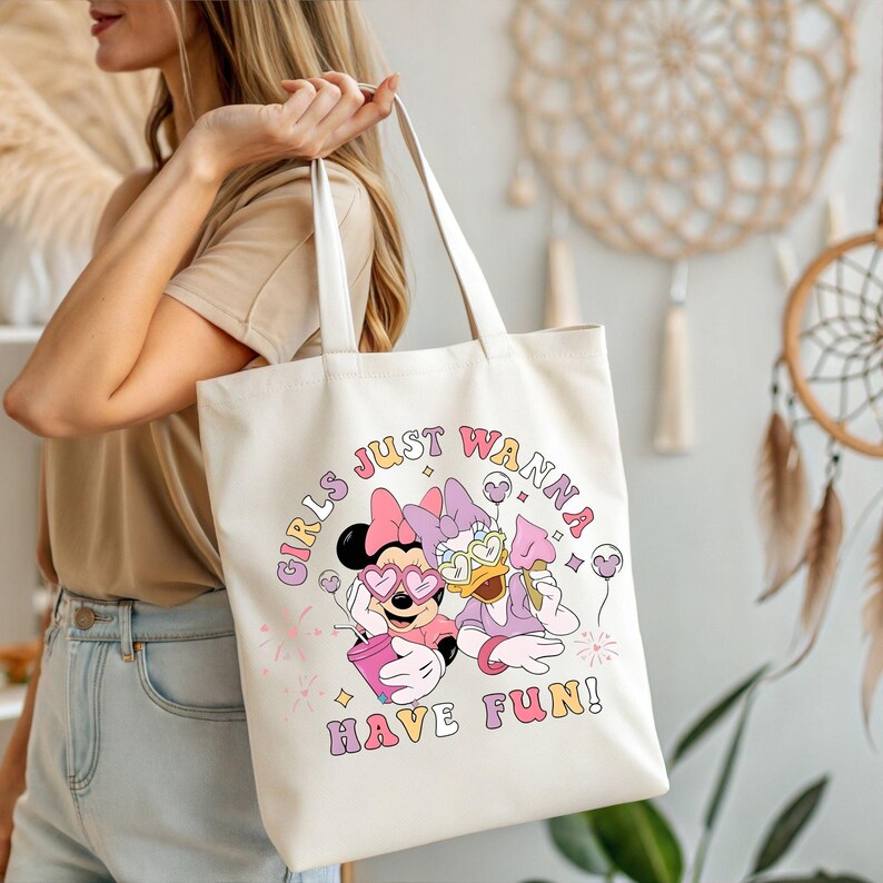 Op de afbeelding: Cr&egrave;mekleurige draagtas met een afbeelding van Minnie Mouse en Daisy Duck met zonnebrillen en ijs. De tekst "GIRLS JUST WANNA HAVE FUN!" is gedrukt in een retro lettertype. De tas heeft witte handvatten.