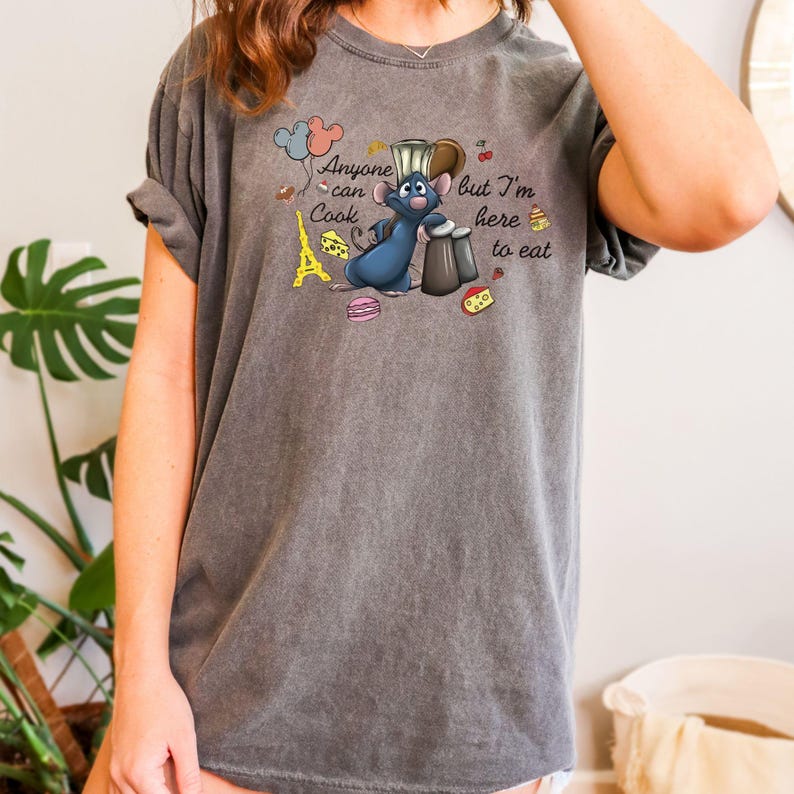 May include: Dark gray t-shirt featuring a cartoon illustration of a blue rat chef with the text "Anyone can cook but I'm here to eat." The design includes a miniature Eiffel Tower, cheese, and macarons.