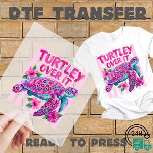 May include: A white t-shirt and a transparent DTF transfer sheet, both featuring a colorful sea turtle with a leopard print shell, pink hibiscus flowers, and the text "TURTLEY OVER IT" in bold pink letters against a light pink striped background.