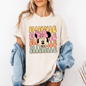 Może przedstawiać: Kremowy t-shirt z retro wzorem. Słowo "MINNIE" jest ułożone pionowo w kolorowych literach bąbelkowych. Twarz Myszki Minnie, z r&oacute;żową kokardą, jest umieszczona pośrodku. Koszulka ma kr&oacute;tkie rękawy.