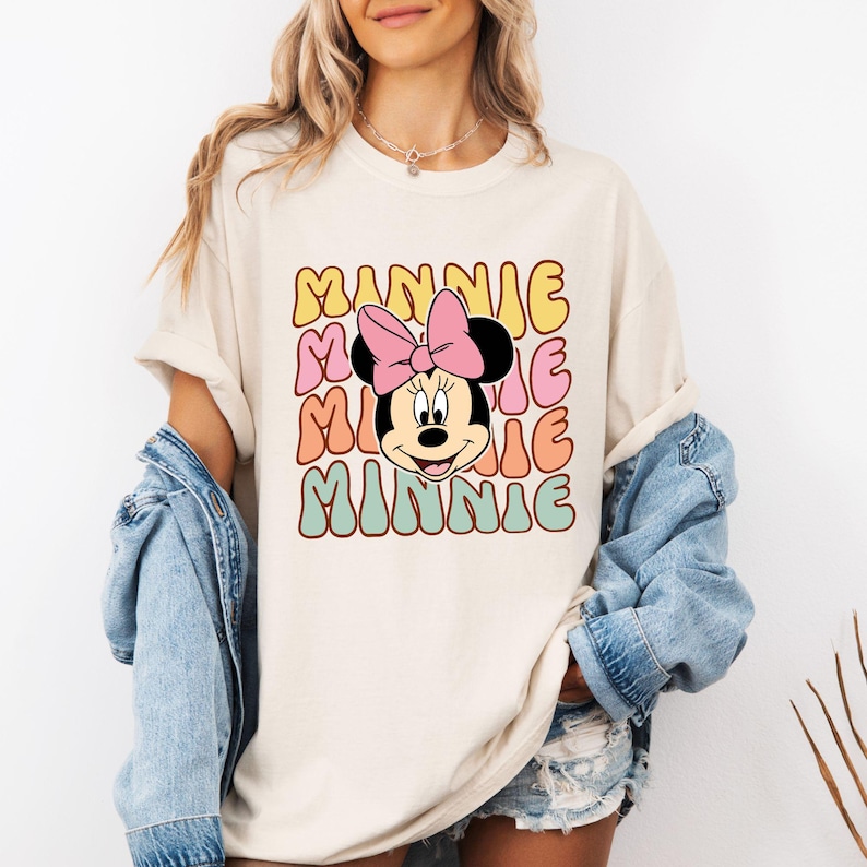 Może przedstawiać: Kremowy t-shirt z retro wzorem. Słowo "MINNIE" jest ułożone pionowo w kolorowych literach bąbelkowych. Twarz Myszki Minnie, z r&oacute;żową kokardą, jest umieszczona pośrodku. Koszulka ma kr&oacute;tkie rękawy.